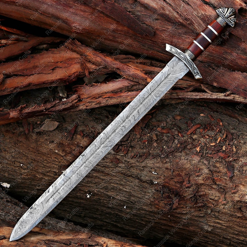 May include: A long, ornate sword with a patterned steel blade and a wooden handle. The handle is dark brown with silver accents. The sword is lying on a bed of brown wood.