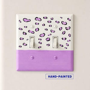 May include: A double light switch wall plate featuring a white top half with a purple and black leopard print pattern and a solid purple bottom half. The plate includes two white toggle switches and four screws. The text HAND-PAINTED is visible.