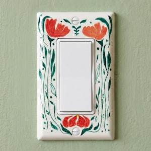 Op de afbeelding: Een rechthoekige witte lichtschakelaar-afdekplaat met een handgeschilderd botanisch ontwerp. De rand toont levendige rode en oranje klaproosachtige bloemen met groene stengels en bladeren, bevestigd met twee zichtbare zilveren schroeven.