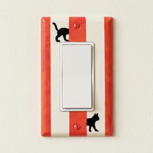 Può includere: Una copertura decorativa per interruttore della luce con un motivo a righe rosse e bianche. La copertura presenta un interruttore bianco e due sagome di gatti neri. È montata su un muro bianco.