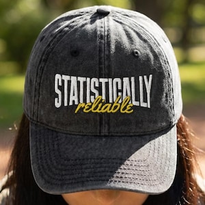 May include: A dark gray baseball cap with the words "STATISTICALLY reliable" embroidered in white and yellow. The cap is worn by a person, with the focus on the hat's design and text.