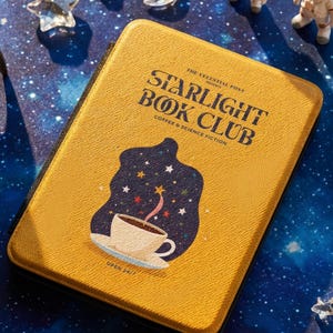 Pode incluir: Uma caixa amarela em forma de livro com o texto "Starlight Book Club" e um desenho de xícara de café. A caixa é cercada por estatuetas de astronautas, pingentes de estrelas e luas e um foguete em um fundo estrelado. O tema é café e ficção científica.