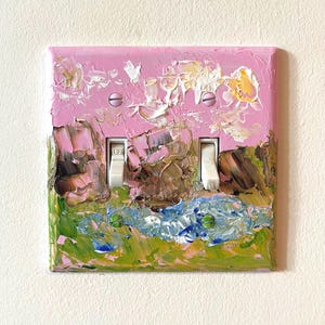 May include: A double light switch wall plate featuring a thick, textured impasto oil painting. The artwork displays a landscape with a pink sky, brown rock formations, and a blue and green grassy foreground. Two white toggle switches are centred.