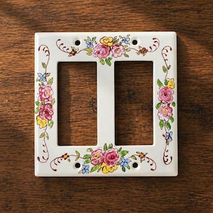 May include: A white double light switch plate featuring a hand-painted floral pattern with pink, yellow, and blue flowers, green leaves, and brown swirling vines. The rectangular cover has four screw holes for mounting on a wall.