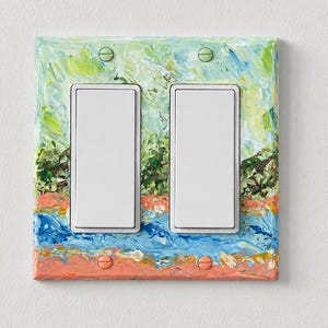 May include: A double light switch wall plate featuring a textured, hand-painted design with thick brushstrokes in shades of light green, dark green, blue, and peach. The plate includes two white rectangular rocker switches and four matching screws.