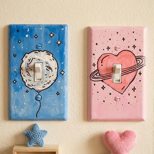 May include: Two decorative light switch covers. One is blue with a moon design, the other is pink with a heart and planet design. Both have white switches and are decorated with stars.