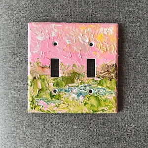 May include: A double light switch plate featuring a thick, textured impasto oil painting of a landscape. The upper portion displays a pink and yellow sky, while the lower section depicts green fields and a blue stream with visible brushstrokes.
