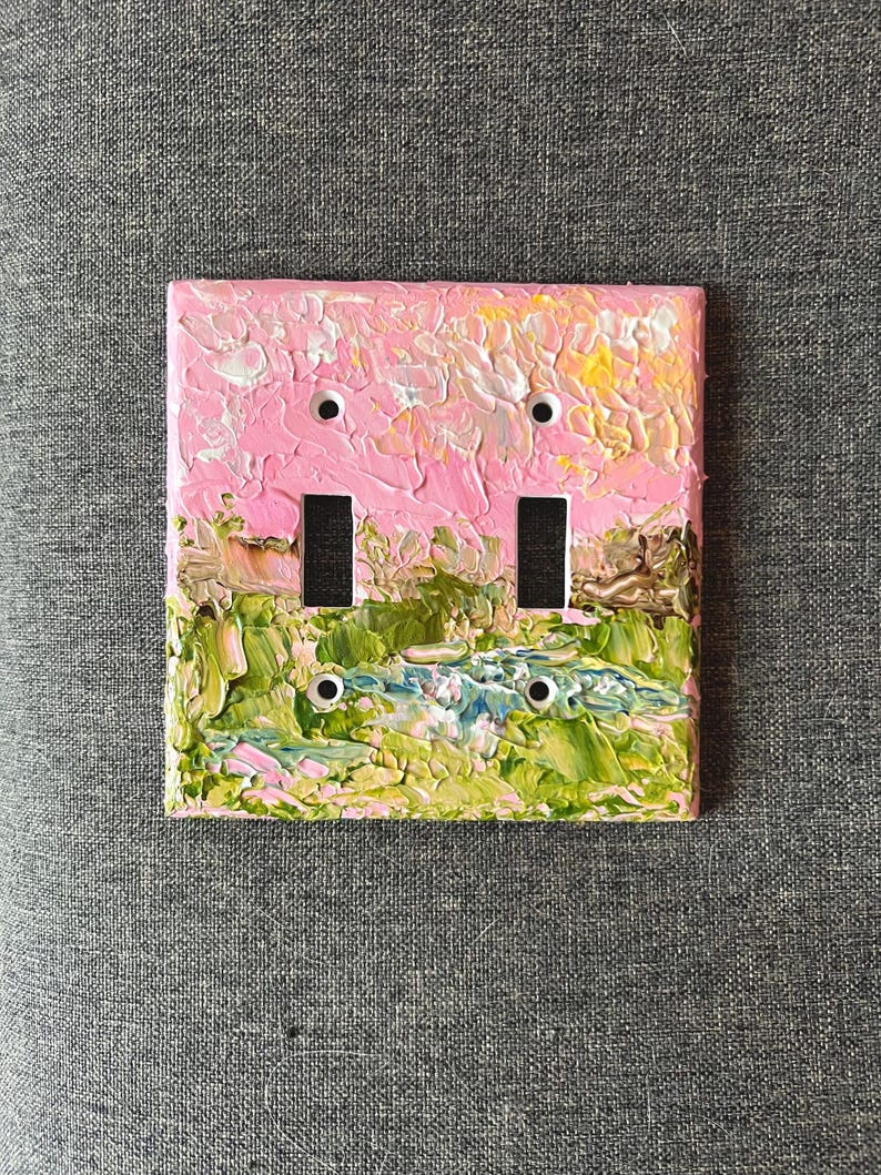 May include: A double light switch plate featuring a thick, textured impasto oil painting of a landscape. The upper portion displays a pink and yellow sky, while the lower section depicts green fields and a blue stream with visible brushstrokes.