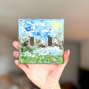 May include: A square double light switch wall plate featuring an impasto-style oil painting of a landscape. The artwork displays a bright blue sky with a yellow sun, green grassy fields, and textured brown and grey mountain peaks.