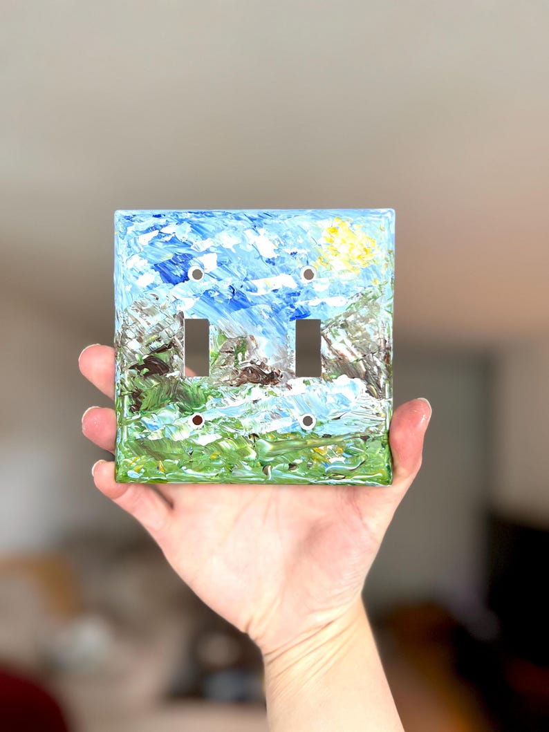 May include: A square double light switch wall plate featuring an impasto-style oil painting of a landscape. The artwork displays a bright blue sky with a yellow sun, green grassy fields, and textured brown and grey mountain peaks.