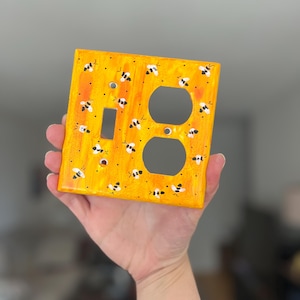Puede incluir: Una placa de pared rectangular con un fondo jaspeado en amarillo y naranja vibrante con abejas pintadas a mano en blanco y negro dispersas por la superficie. La placa incluye un recorte rectangular y dos recortes circulares para enchufes.