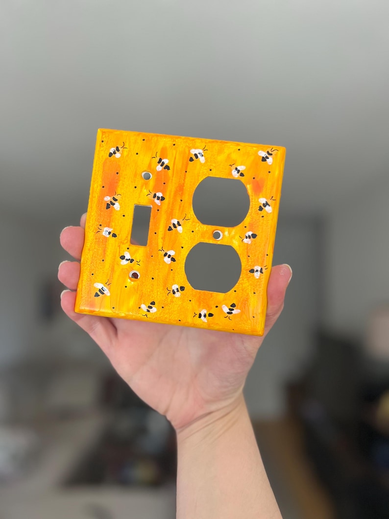 Puede incluir: Una placa de pared rectangular con un fondo jaspeado en amarillo y naranja vibrante con abejas pintadas a mano en blanco y negro dispersas por la superficie. La placa incluye un recorte rectangular y dos recortes circulares para enchufes.