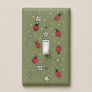May include: A rectangular light olive green light switch plate featuring a hand-painted pattern of red ladybirds with black spots and small white flowers with yellow centres, accented by tiny white dots and secured with two flat-head screws.