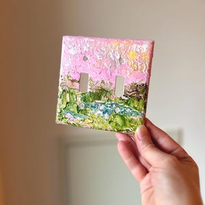 May include: A double light switch plate cover featuring a thick, textured impasto oil painting of a landscape. The design displays a vibrant pink sky above a lush green field with a blue stream, applied with visible, raised brushstrokes.