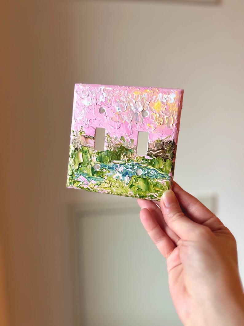 May include: A double light switch plate cover featuring a thick, textured impasto oil painting of a landscape. The design displays a vibrant pink sky above a lush green field with a blue stream, applied with visible, raised brushstrokes.