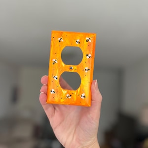Puede incluir: Una placa de pared rectangular de color naranja para toma de corriente con un dise&ntilde;o pintado a mano de peque&ntilde;as abejas negras y blancas dispersas por la superficie. La placa tiene dos aberturas circulares y un orificio central.