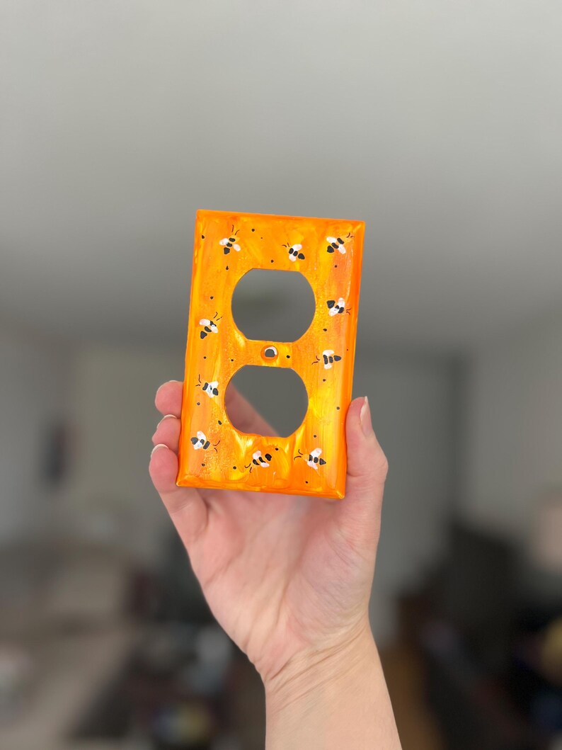 Puede incluir: Una placa de pared rectangular de color naranja para toma de corriente con un dise&ntilde;o pintado a mano de peque&ntilde;as abejas negras y blancas dispersas por la superficie. La placa tiene dos aberturas circulares y un orificio central.