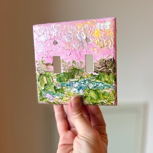 May include: A square double light switch wall plate featuring a thick, textured impasto oil painting. The design depicts a landscape with a vibrant pink sky above green foliage and a blue water element, applied with visible, raised brushstrokes.