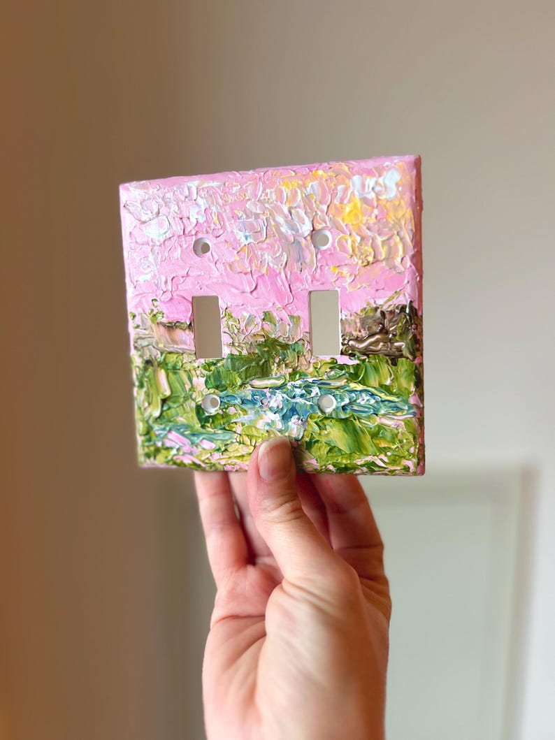 May include: A square double light switch wall plate featuring a thick, textured impasto oil painting. The design depicts a landscape with a vibrant pink sky above green foliage and a blue water element, applied with visible, raised brushstrokes.