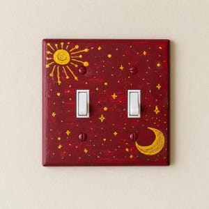 May include: A decorative light switch cover with a celestial design. The maroon cover is painted with a yellow sun, moon, and stars. Two white light switches are in the centre.