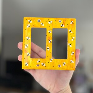 Puede incluir: Una placa de pared rectangular para interruptor doble con un fondo amarillo brillante y un patr&oacute;n pintado a mano de peque&ntilde;as abejas blancas y negras dispersas por la superficie. La placa incluye cuatro orificios circulares.