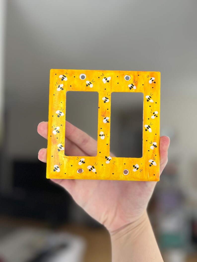 Puede incluir: Una placa de pared rectangular para interruptor doble con un fondo amarillo brillante y un patr&oacute;n pintado a mano de peque&ntilde;as abejas blancas y negras dispersas por la superficie. La placa incluye cuatro orificios circulares.