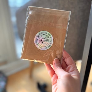Puede incluir: Un sobre de papel marr&oacute;n sellado en una funda de pl&aacute;stico transparente, con una pegatina hologr&aacute;fica circular que muestra una ilustraci&oacute;n de un planeta y el texto The Celestial Post Your Interstellar Postal Office en tinta negra.