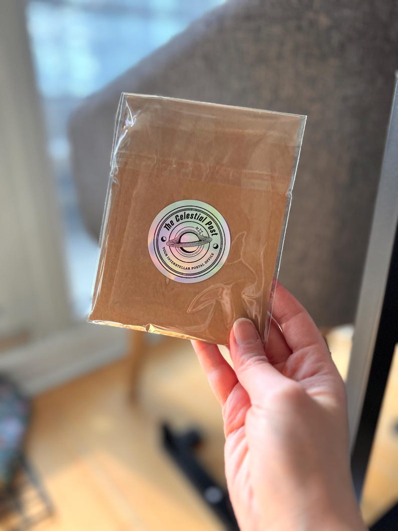 Puede incluir: Un sobre de papel marr&oacute;n sellado en una funda de pl&aacute;stico transparente, con una pegatina hologr&aacute;fica circular que muestra una ilustraci&oacute;n de un planeta y el texto The Celestial Post Your Interstellar Postal Office en tinta negra.