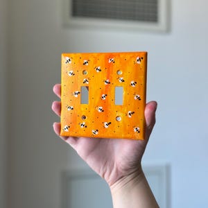 Puede incluir: Una placa de pared rectangular para interruptor doble con un fondo pintado en naranja y amarillo vibrante, decorada con un patr&oacute;n repetitivo de peque&ntilde;as abejas pintadas a mano en blanco y negro sobre toda la superficie.