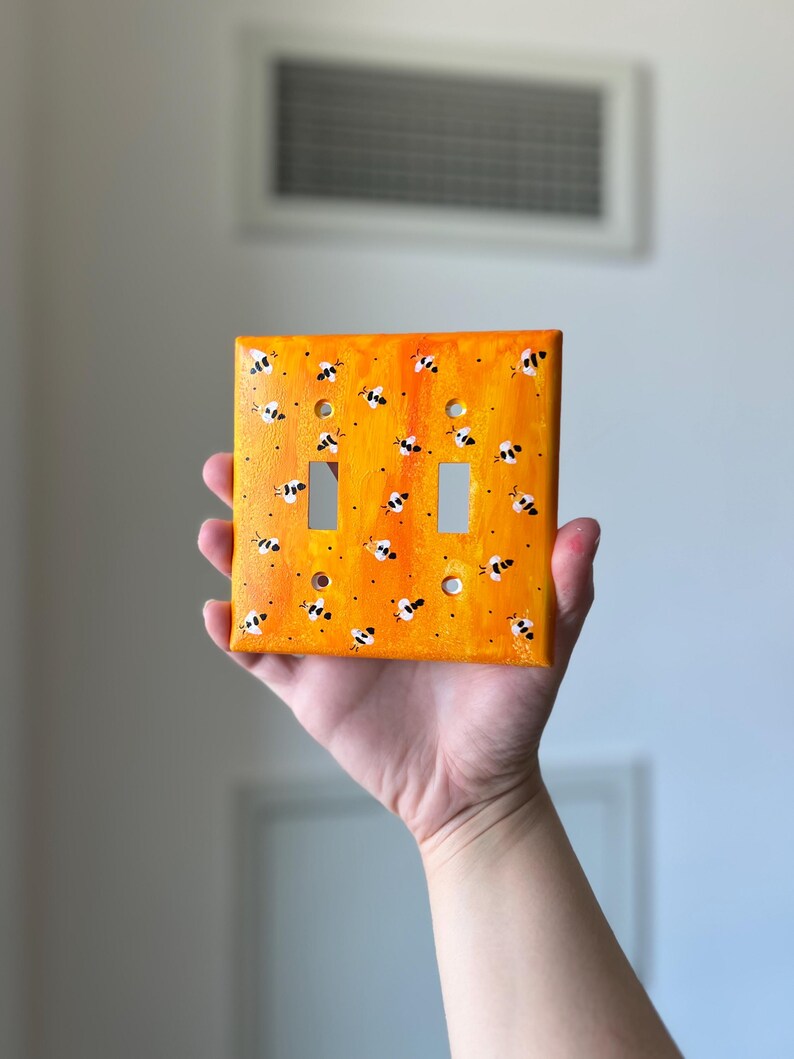 Puede incluir: Una placa de pared rectangular para interruptor doble con un fondo pintado en naranja y amarillo vibrante, decorada con un patr&oacute;n repetitivo de peque&ntilde;as abejas pintadas a mano en blanco y negro sobre toda la superficie.
