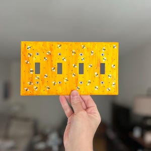 Puede incluir: Una placa rectangular amarilla para interruptor de luz con un dise&ntilde;o pintado a mano de numerosas abejas peque&ntilde;as en blanco y negro dispersas por la superficie. La placa incluye cuatro aberturas rectangulares verticales.