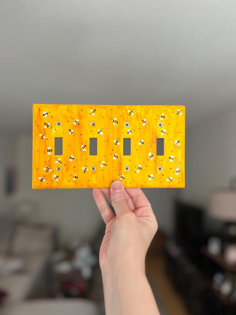 Puede incluir: Una placa rectangular amarilla para interruptor de luz con un dise&ntilde;o pintado a mano de numerosas abejas peque&ntilde;as en blanco y negro dispersas por la superficie. La placa incluye cuatro aberturas rectangulares verticales.