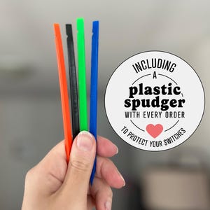 May include: Four thin plastic spudger tools in orange, black, green, and blue. A circular white sticker features the black text: INCLUDING A plastic spudger WITH EVERY ORDER TO PROTECT YOUR SWITCHES, with a small red heart icon below the text.