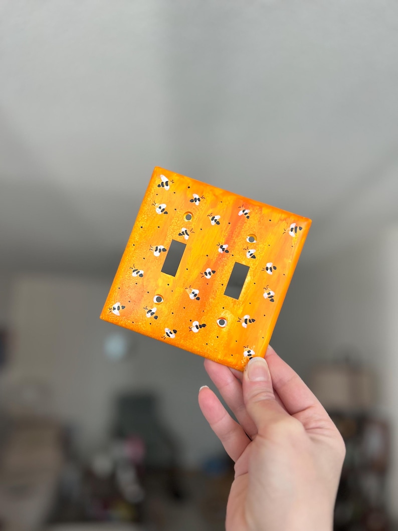 Puede incluir: Una placa de cubierta para interruptor doble rectangular con un fondo de degradado naranja y amarillo vibrante y un patr&oacute;n pintado a mano de peque&ntilde;as abejas negras y blancas dispersas por la superficie, incluyendo dos aberturas rectangulares.