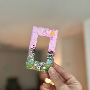 May include: A rectangular light switch wall plate featuring a textured, hand-painted design. The top section is primarily light pink with small yellow accents, while the bottom section transitions into a blend of green, black, and pink brushstrokes.