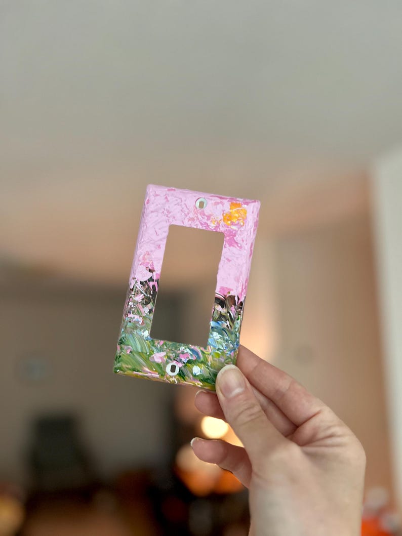 May include: A rectangular light switch wall plate featuring a textured, hand-painted design. The top section is primarily light pink with small yellow accents, while the bottom section transitions into a blend of green, black, and pink brushstrokes.