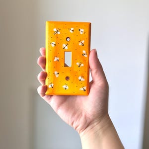 Puede incluir: Una placa rectangular para interruptor de luz pintada en un tono amarillo vibrante. La superficie presenta un patr&oacute;n repetitivo de peque&ntilde;as abejas blancas y negras pintadas a mano, dispersas por el frente junto a diminutos puntos negros.