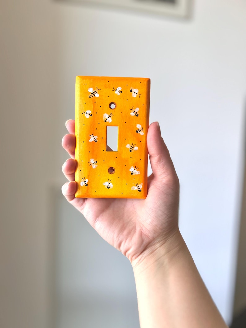 Puede incluir: Una placa rectangular para interruptor de luz pintada en un tono amarillo vibrante. La superficie presenta un patr&oacute;n repetitivo de peque&ntilde;as abejas blancas y negras pintadas a mano, dispersas por el frente junto a diminutos puntos negros.