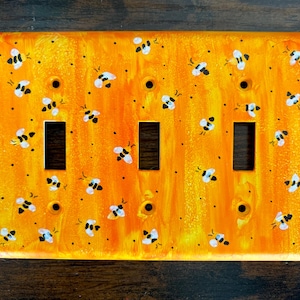 Puede incluir: Una placa de pared rectangular para triple interruptor con un fondo jaspeado en tonos naranja y amarillo vibrante, decorada con un patr&oacute;n repetitivo de peque&ntilde;as abejas pintadas a mano en blanco y negro y puntos negros dispersos.
