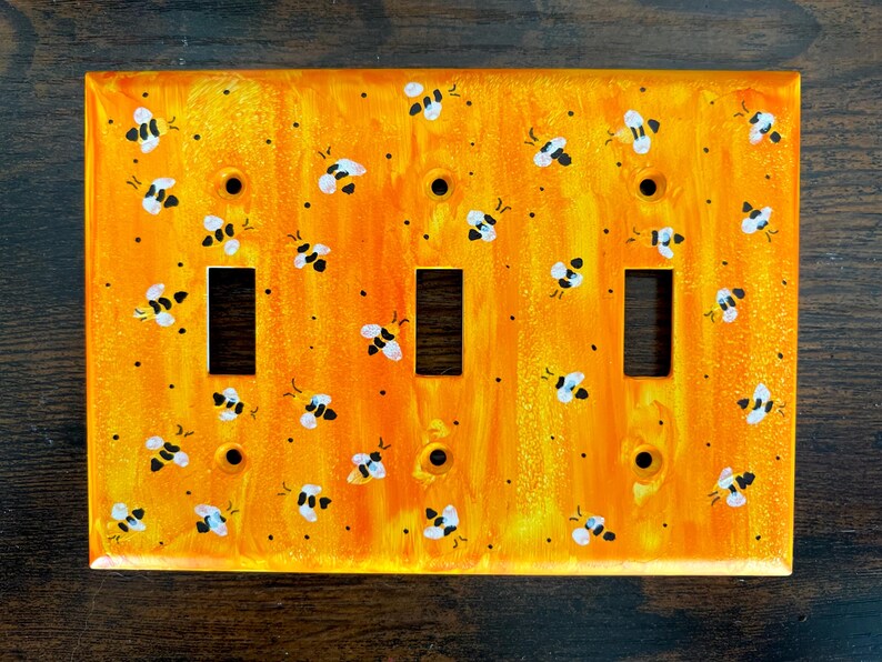 Puede incluir: Una placa de pared rectangular para triple interruptor con un fondo jaspeado en tonos naranja y amarillo vibrante, decorada con un patr&oacute;n repetitivo de peque&ntilde;as abejas pintadas a mano en blanco y negro y puntos negros dispersos.