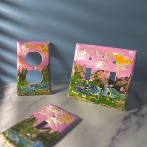 May include: Three rectangular light switch plates featuring a hand-painted landscape design with thick, textured brushstrokes. The artwork displays a pink sky with yellow clouds above green hills and blue water, applied to standard electrical wall covers.