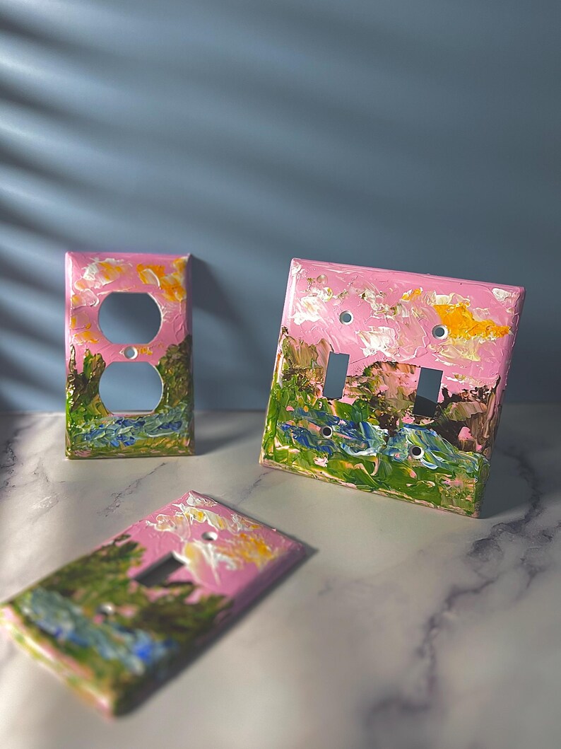 May include: Three rectangular light switch plates featuring a hand-painted landscape design with thick, textured brushstrokes. The artwork displays a pink sky with yellow clouds above green hills and blue water, applied to standard electrical wall covers.