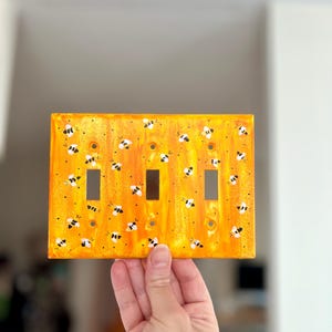 Puede incluir: Una placa de pared rectangular para triple interruptor de luz con un fondo jaspeado en tonos naranja y amarillo vibrantes. La superficie est&aacute; decorada con numerosas abejas peque&ntilde;as pintadas a mano en blanco y negro por toda la placa.
