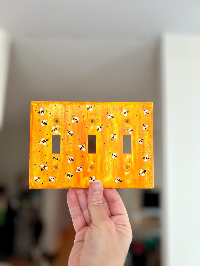 Puede incluir: Una placa de pared rectangular para triple interruptor de luz con un fondo jaspeado en tonos naranja y amarillo vibrantes. La superficie est&aacute; decorada con numerosas abejas peque&ntilde;as pintadas a mano en blanco y negro por toda la placa.