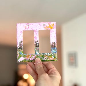 May include: A rectangular double light switch cover plate featuring a hand-painted design with a light pink background and textured strokes of green, blue, and yellow paint along the bottom edge. The plate has two vertical rectangular openings.