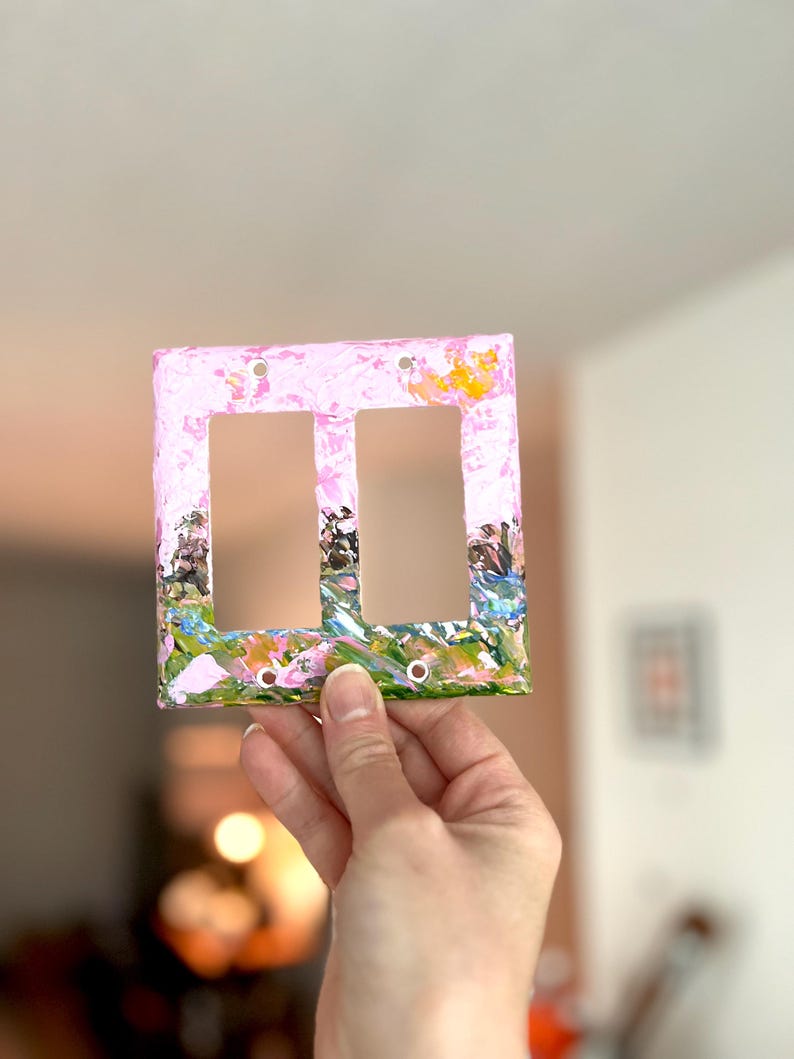 May include: A rectangular double light switch cover plate featuring a hand-painted design with a light pink background and textured strokes of green, blue, and yellow paint along the bottom edge. The plate has two vertical rectangular openings.