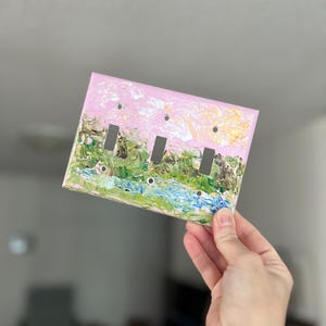 May include: A rectangular triple light switch cover plate featuring an impasto-style painted landscape. The design displays a light pink sky above a textured green forest and a blue stream, with three vertical rectangular cutouts for switches.