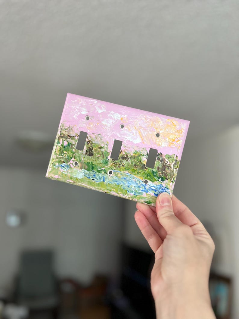 May include: A rectangular triple light switch cover plate featuring an impasto-style painted landscape. The design displays a light pink sky above a textured green forest and a blue stream, with three vertical rectangular cutouts for switches.