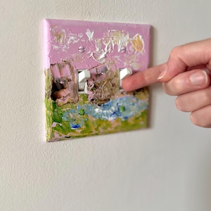 May include: A double light switch plate featuring a textured, impasto-style oil painting with a pink sky, green grass, and blue water. The rectangular cover has two white toggle switches and is secured with two pink-painted metal screws.