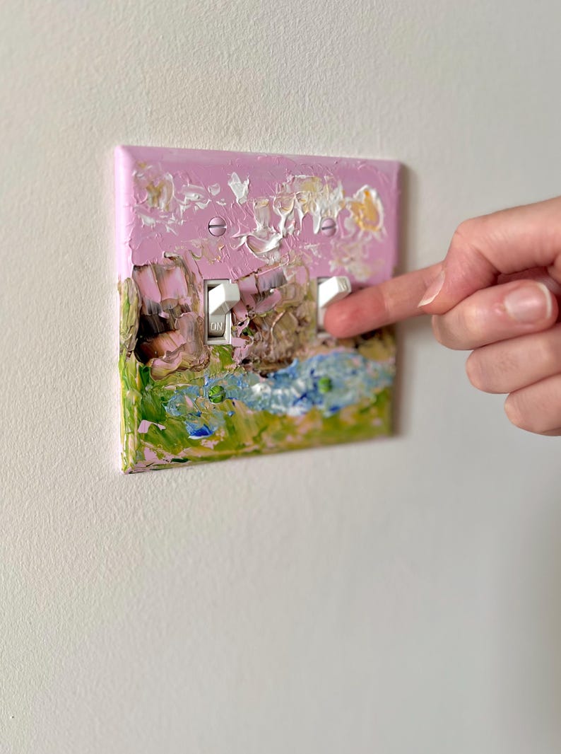 May include: A double light switch plate featuring a textured, impasto-style oil painting with a pink sky, green grass, and blue water. The rectangular cover has two white toggle switches and is secured with two pink-painted metal screws.